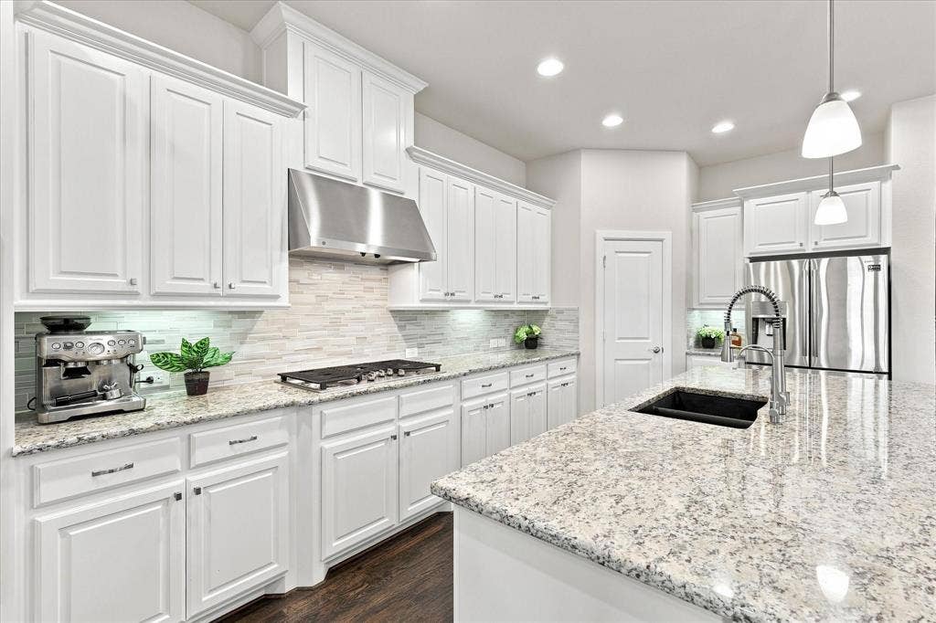 White cabinetry with crown molding, light-toned countertops, stainless steel appliances, a center island with a sink, and pendant lighting