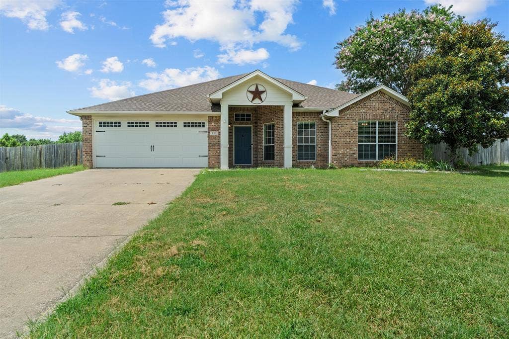 833 Greenwood Circle, Lindale, TX 75771 Charming one-story brick residence featuring a two-car garage with paneled doors, arched entryway with decorative star accent, multiple large windows with black trim, and a spacious front lawn
