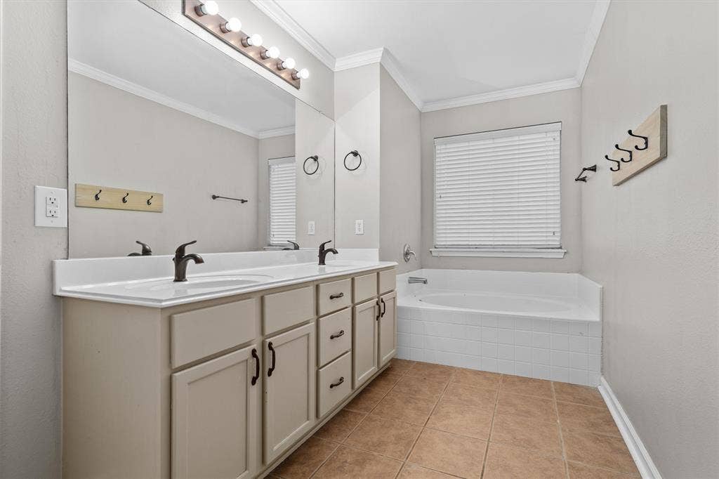 833 Greenwood Circle, Lindale, TX 75771 Primary bathroom featuring a dual-sink vanity with wood-finish cabinetry, white countertop, and matte black faucets; framed mirror with integrated lighting; soaking tub with tile surround; tile flooring; and a window with plantation shutters