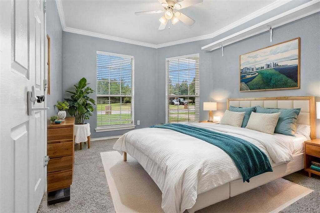 833 Greenwood Circle, Lindale, TX 75771 Serene primary bedroom suite featuring dual glass doors opening to outdoor landscape, soft blue-gray wall finishes, wood-finish nightstands with integrated lighting, ceiling fan with light fixture, and plush bedding in complementary tones