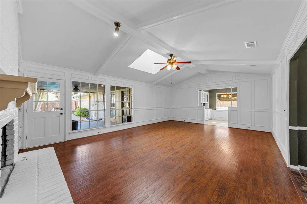 1116 Windsong Trail, Richardson, TX 75081 Vaulted ceiling with exposed beams, wood-finish flooring, white painted brick fireplace, paneled accent walls, and a ceiling fan with integrated lighting