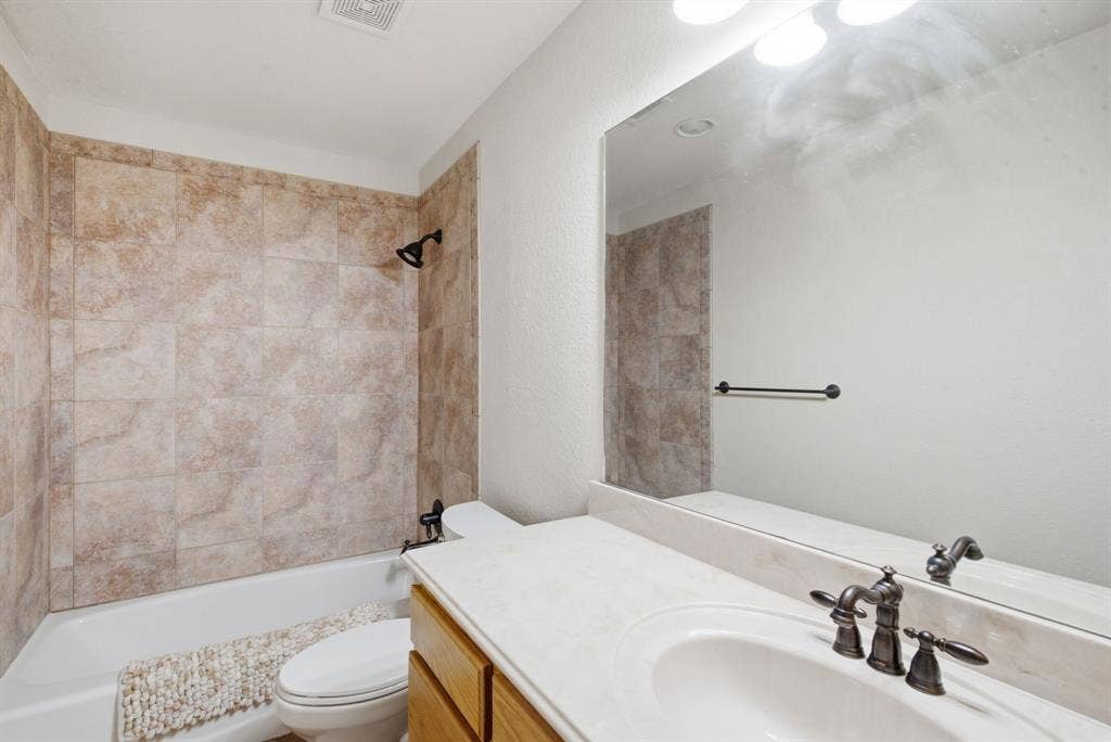600 N Avalon Court, Granbury, TX 76048 Upstairs bathroom featuring a neutral stone-tile tub surround, light-toned countertop with integrated basin, dark finish plumbing fixtures, large vanity mirror, and wood-finish cabinetry