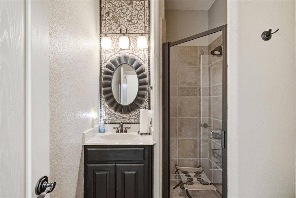 600 N Avalon Court, Granbury, TX 76048 Downstairs bathroom featuring a dark-finish vanity with a light-toned countertop, a decorative tiled wall accent, a scalloped-edge mirror, and a tiled shower enclosure with a dark-finish frame