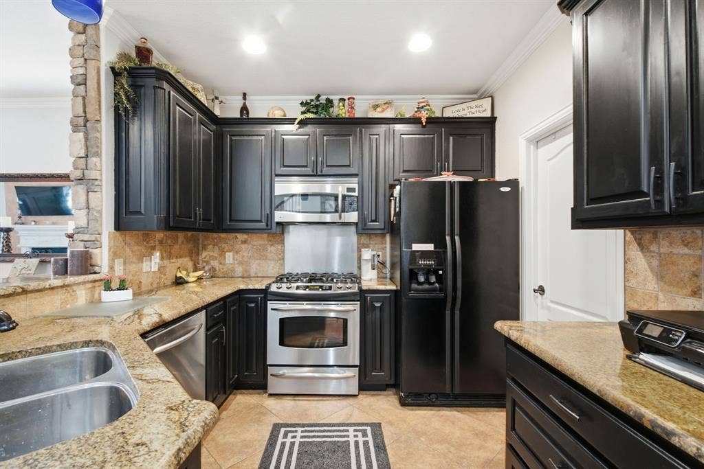 600 N Avalon Court, Granbury, TX 76048 Kitchen featuring dark cabinetry, stone-finish countertops, tile backsplash, stainless steel appliances, and a stacked-stone pillar accent
