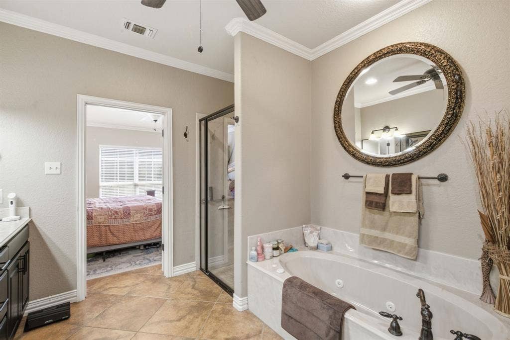 600 N Avalon Court, Granbury, TX 76048 Jetted soaking tub with a stone-surround deck, glass-enclosed shower, dark-finish cabinetry, ornate circular wall mirror, and crown molding