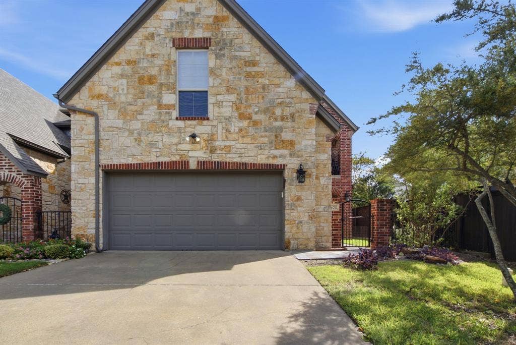 600 N Avalon Court, Granbury, TX 76048 Stone and brick exterior facade featuring a concrete driveway, multi-gabled roofline, metal side-entry gate, and carriage-style outdoor wall lighting