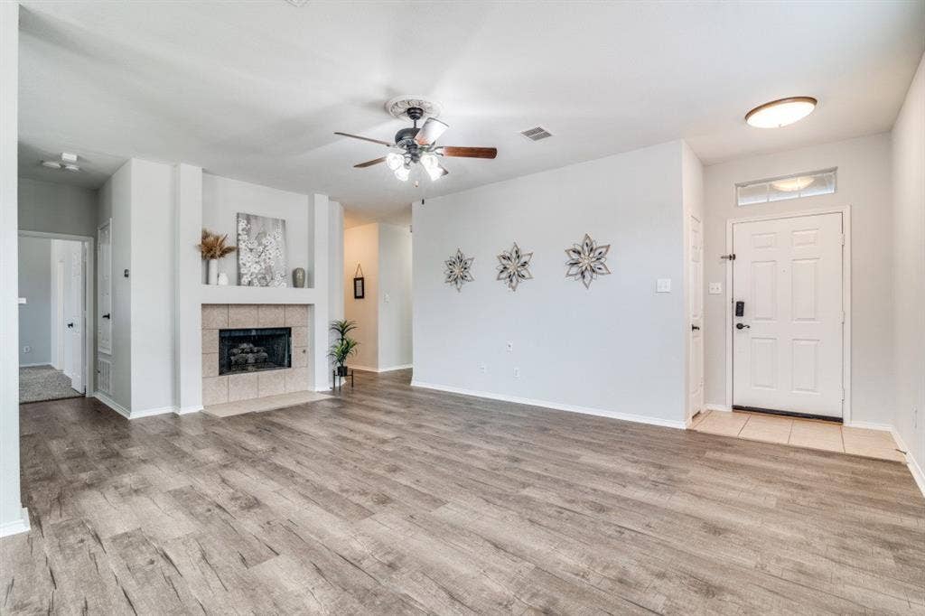 Wood-finish flooring throughout, ceiling fan with integrated lighting, recessed fireplace with tile surround, white entry door with transom window, and flush mount ceiling light fixture