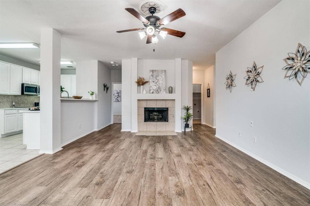 Wood-finish flooring throughout, ceiling fan with light fixture, fireplace with tile surround, and an open concept kitchen with white cabinetry