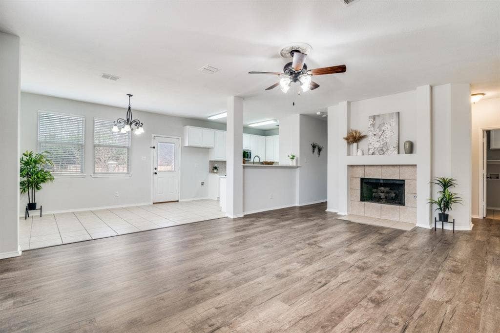 Wood-finish flooring, tiled fireplace with mantel, ceiling fan, white kitchen cabinetry, and pendant light fixture