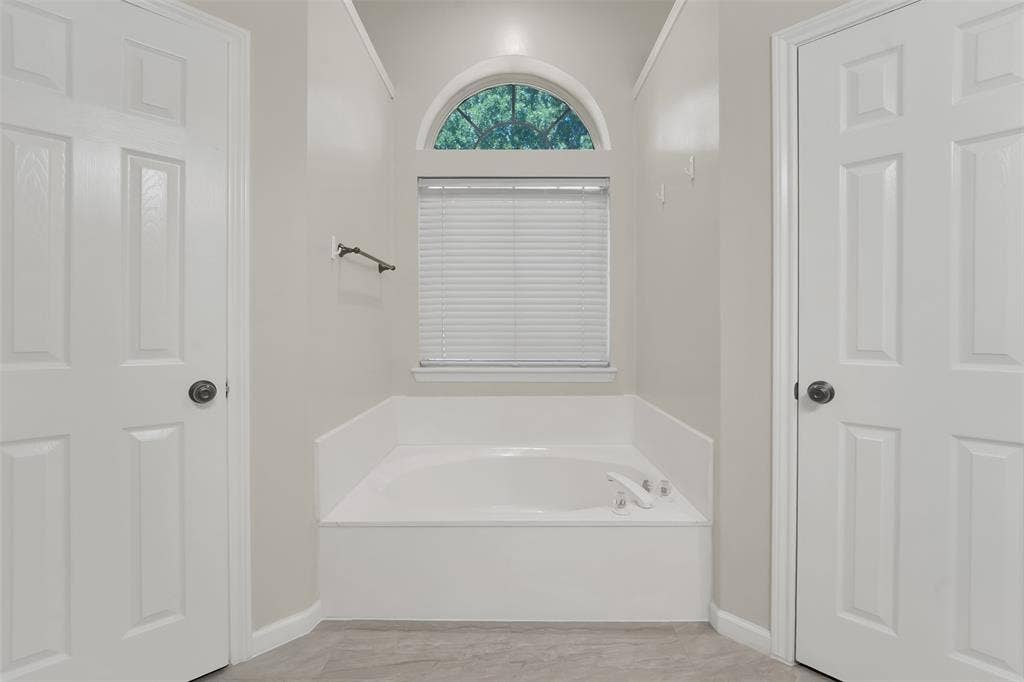 4001 Arrowcrest Lane, Garland, TX 75044 Primary Bathroom featuring a spacious soaking tub with integrated controls, ceramic tile surround, neutral finish flooring, dual six-panel closet doors, and a transom window with operable shutters providing natural light