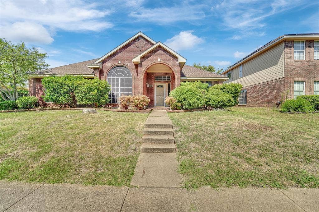 4001 Arrowcrest Lane, Garland, TX 75044 Charming brick residence featuring a welcoming arched entryway, manicured front lawn with mature landscaping, and a concrete walkway leading to the home's entrance