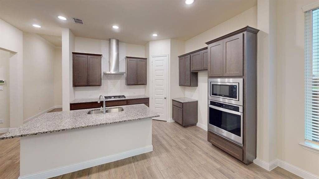 3314 Willena Road, Melissa, TX 75454 Modern kitchen featuring a large center island with waterfall-edge countertop, stainless steel appliances including dual wall ovens and a gas cooktop with overhead hood, dark cabinetry, granite countertops, wood-finish flooring, and abundant recessed lighting throughout the open layout