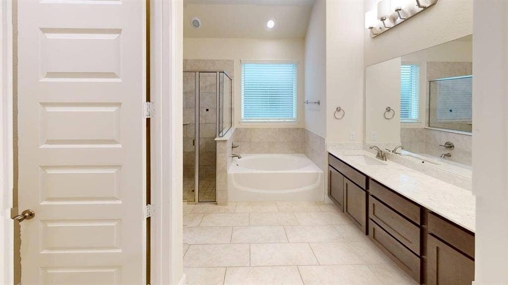 3314 Willena Road, Melissa, TX 75454 Spa-inspired primary bathroom featuring a soaking tub with glass enclosure, dual-sink vanity with wood cabinetry and quartz countertop, large framed mirrors with integrated lighting, tile flooring, and a window with plantation shutters