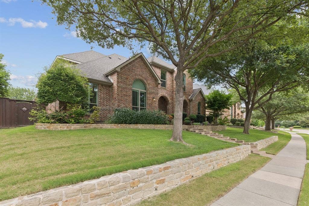 15456 Mountain View Lane, Frisco, TX 75035 Brick facade with arched windows, tiered landscaping featuring stone retaining walls, mature trees, and a concrete sidewalk