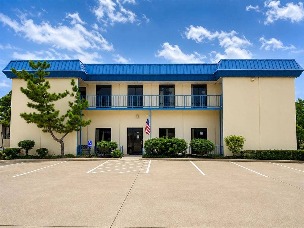 1039 N Interstate 35 Highway N, #E, Carrollton, TX 75006 Two-story commercial building with a blue metal roof, cream-colored exterior siding, and a second-story balcony with blue railings