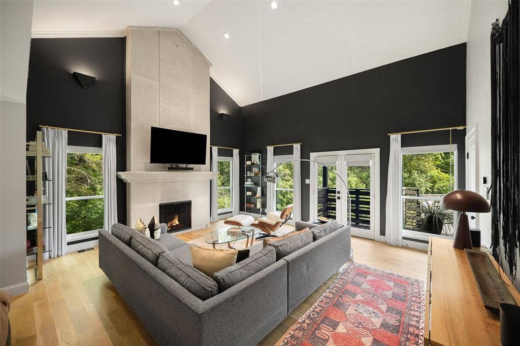 9863 Elmcrest Drive, Dallas, TX 75238 Primary suite living area showcasing a soaring vaulted ceiling, a stone-clad fireplace, light wood-finish flooring, and multiple large windows with access to an exterior balcony.