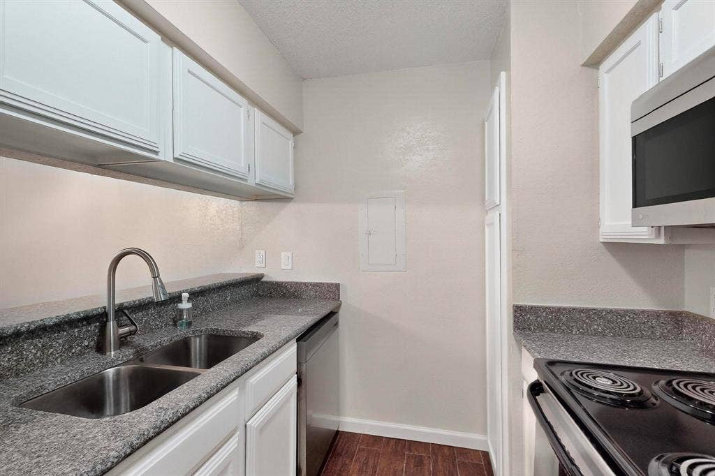 White upper and lower cabinetry, dual basin stainless steel sink with gooseneck faucet, dark speckled countertops, wood-finish flooring, and a stainless steel over-the-range microwave