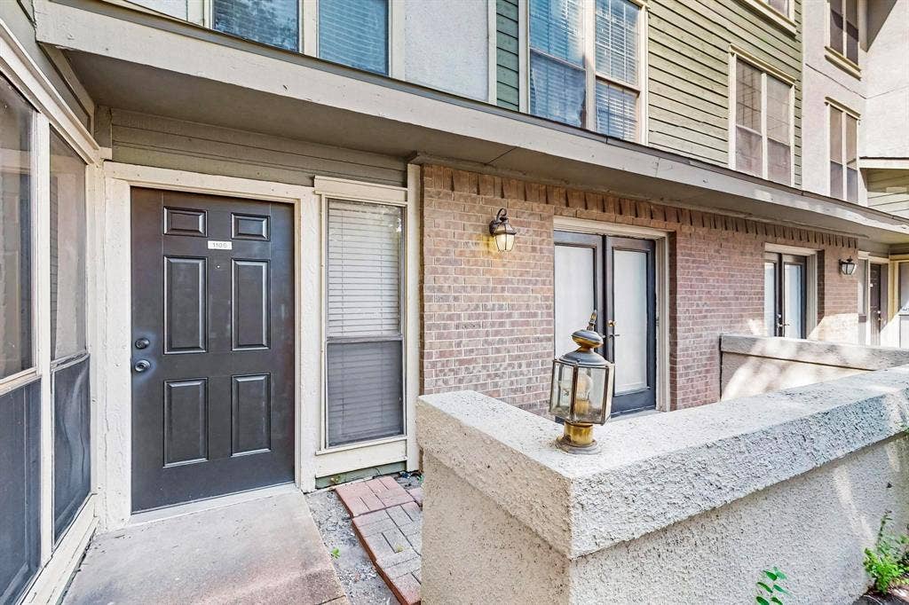 Brick exterior with concrete patio, black paneled entry door, adjacent window, and outdoor lighting fixtures