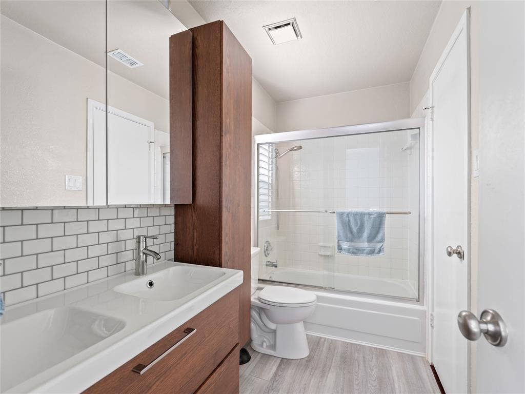 6220 Bentwood Trail, #205, Dallas, TX 75252 Vanity with integrated dual sinks, subway tile backsplash, wood-finish cabinetry, and a shower-tub combination with sliding glass doors