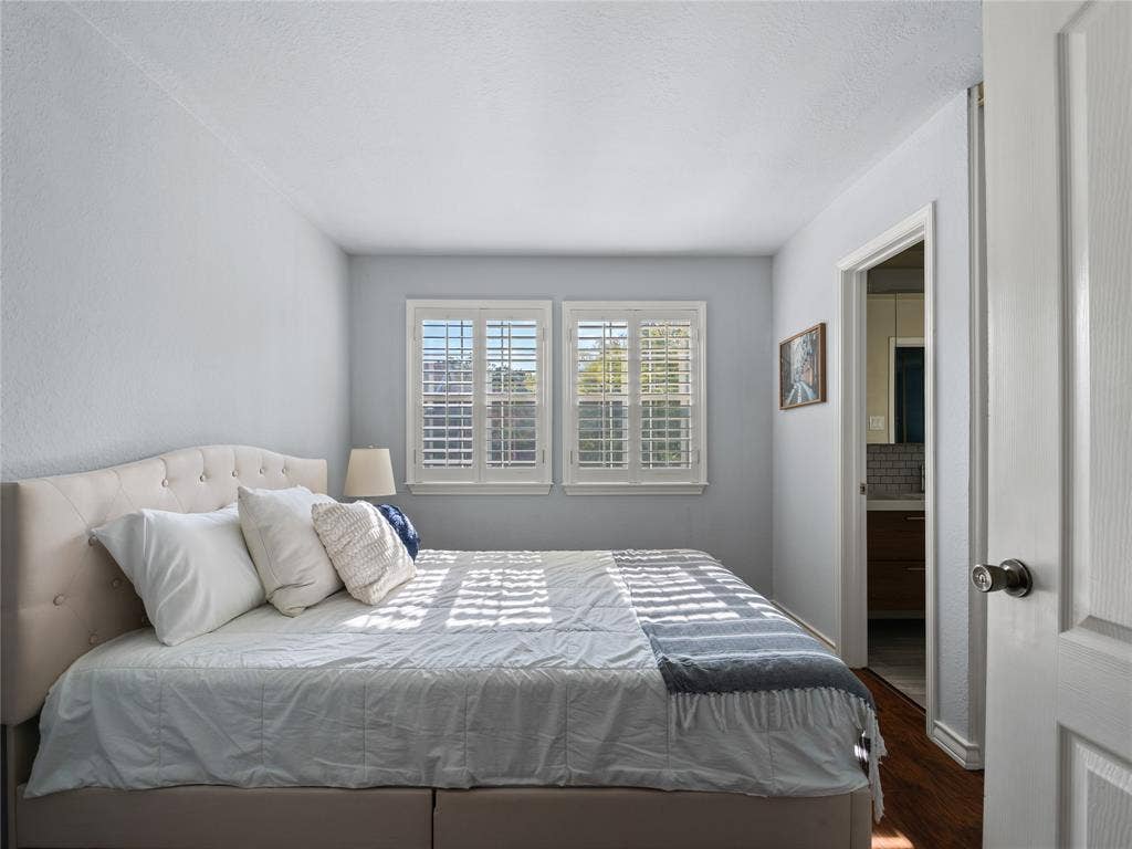 6220 Bentwood Trail, #205, Dallas, TX 75252 Two windows with white plantation shutters, an upholstered tufted headboard, and wood-finish flooring