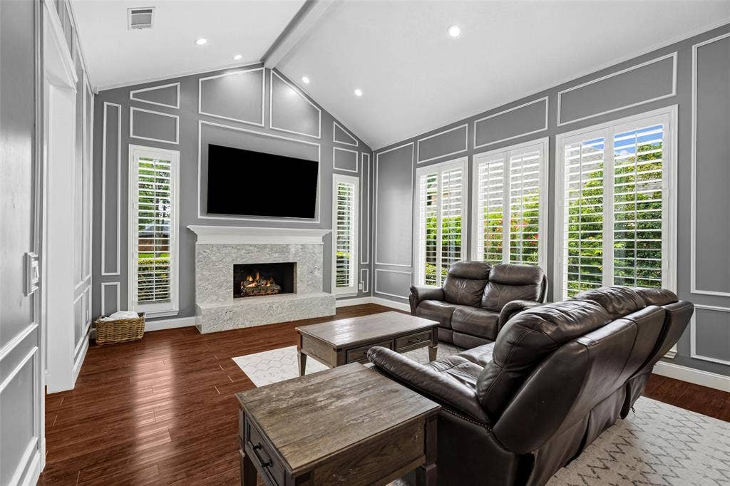 2705 Ithaca Court, Plano, TX 75025 Living area featuring vaulted ceilings, wood-finish flooring, a fireplace with a stone surround, and multiple tall windows with plantation shutters