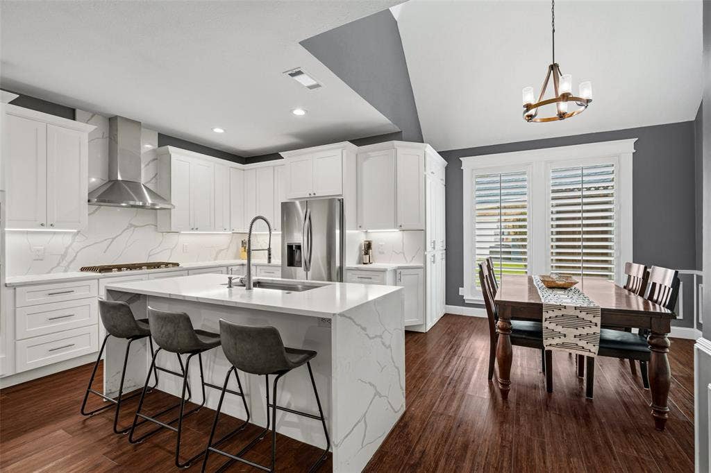 2705 Ithaca Court, Plano, TX 75025 Modern kitchen and dining area featuring white cabinetry, marble-finish island with waterfall edge and seating for three, stainless steel appliances, pendant lighting, wood-finish flooring, and plantation shutters overlooking the dining space with coffered ceiling detail