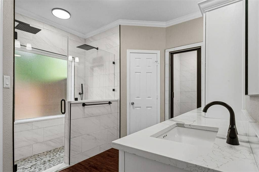 2705 Ithaca Court, Plano, TX 75025 Dual shower heads, glass-enclosed shower with tiled walls and pebble-finish floor, white vanity with integrated sink, black-finish faucet, and wood-finish flooring