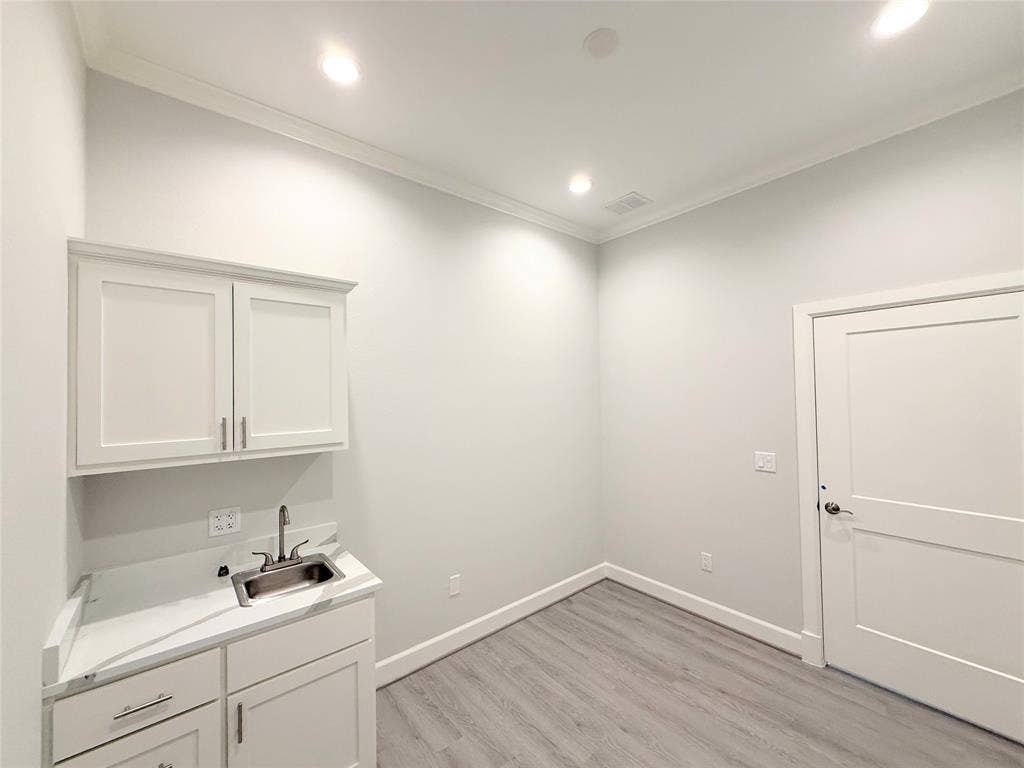2120 Prairie Drive, #901, Prosper, TX 75078 Bright utility room featuring white cabinetry with brushed nickel hardware, a stainless steel sink with single-lever faucet, quartz countertop, wood-finish flooring, recessed lighting, and a white painted entry door