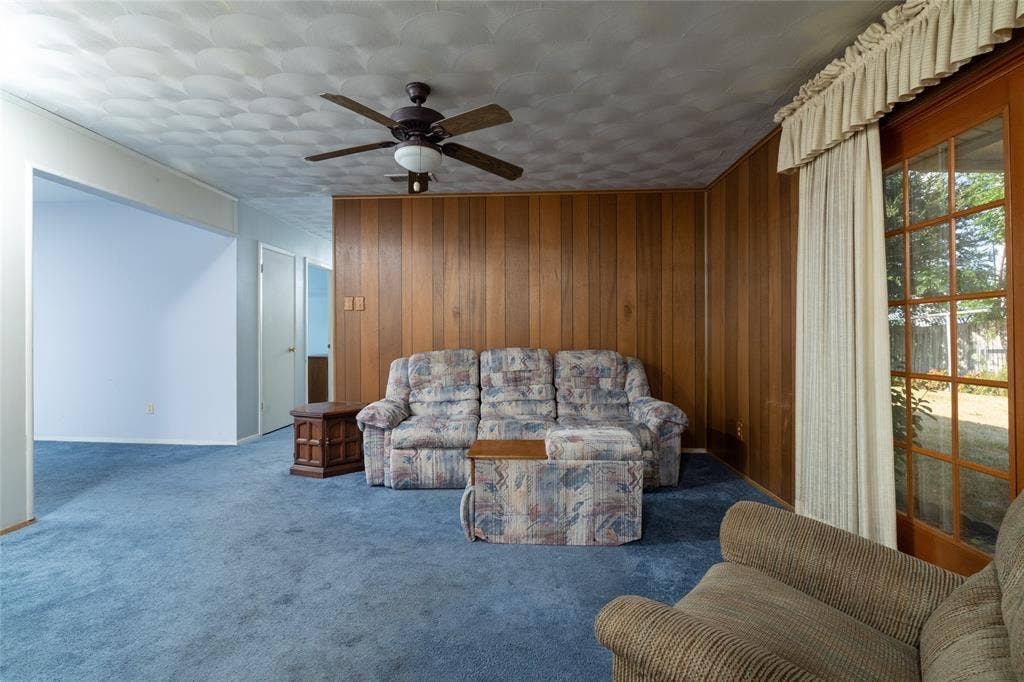 Blue carpeting, wood paneled accent wall, ceiling fan with integrated lighting, and glass-paned exterior door
