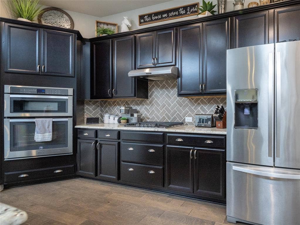 Black cabinetry with silver hardware, light-toned countertops, herringbone pattern tile backsplash, stainless steel double wall oven, and wood-finish flooring