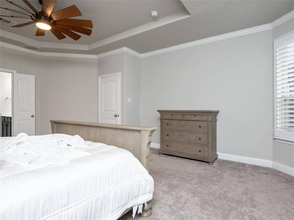Tray ceiling with crown molding, wood-finish ceiling fan, gray carpeting, and window with plantation shutters