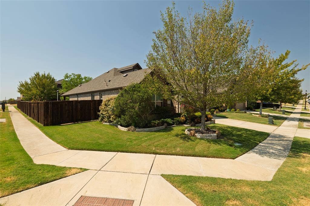Brick exterior with a dark shingle roof, mature trees, landscaped shrubbery, and a wood privacy fence