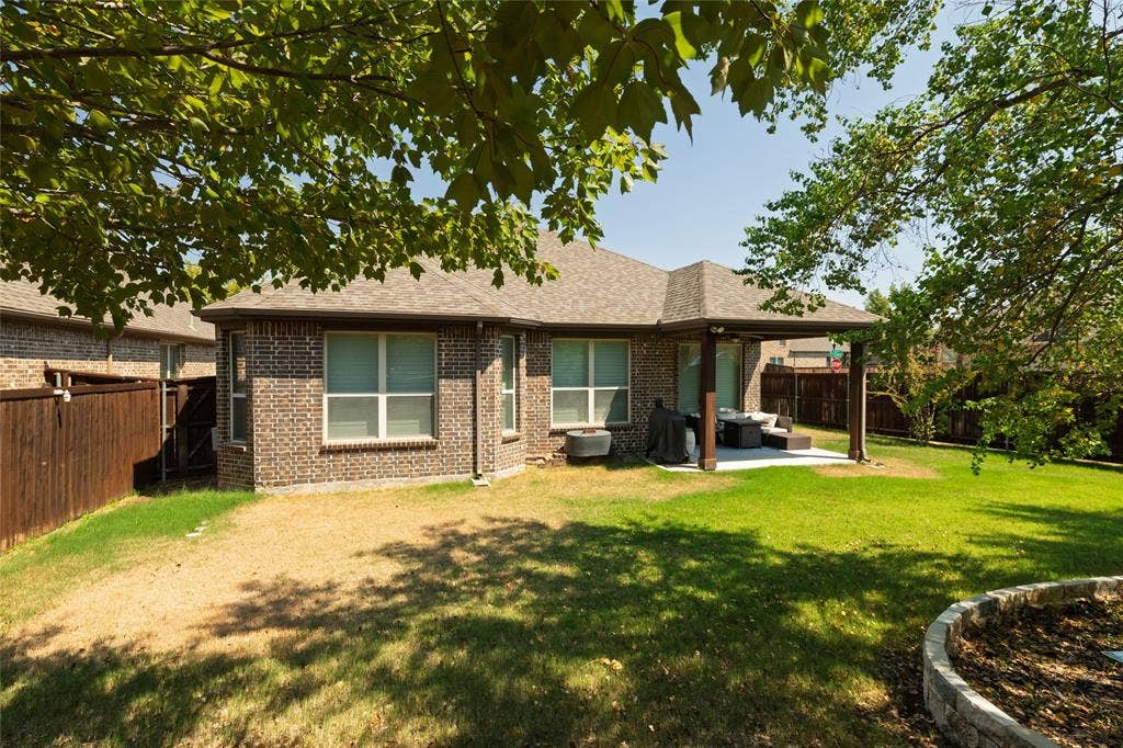 Brick exterior with multiple windows, covered patio, wood fencing, a stone-edged planting area, and mature trees