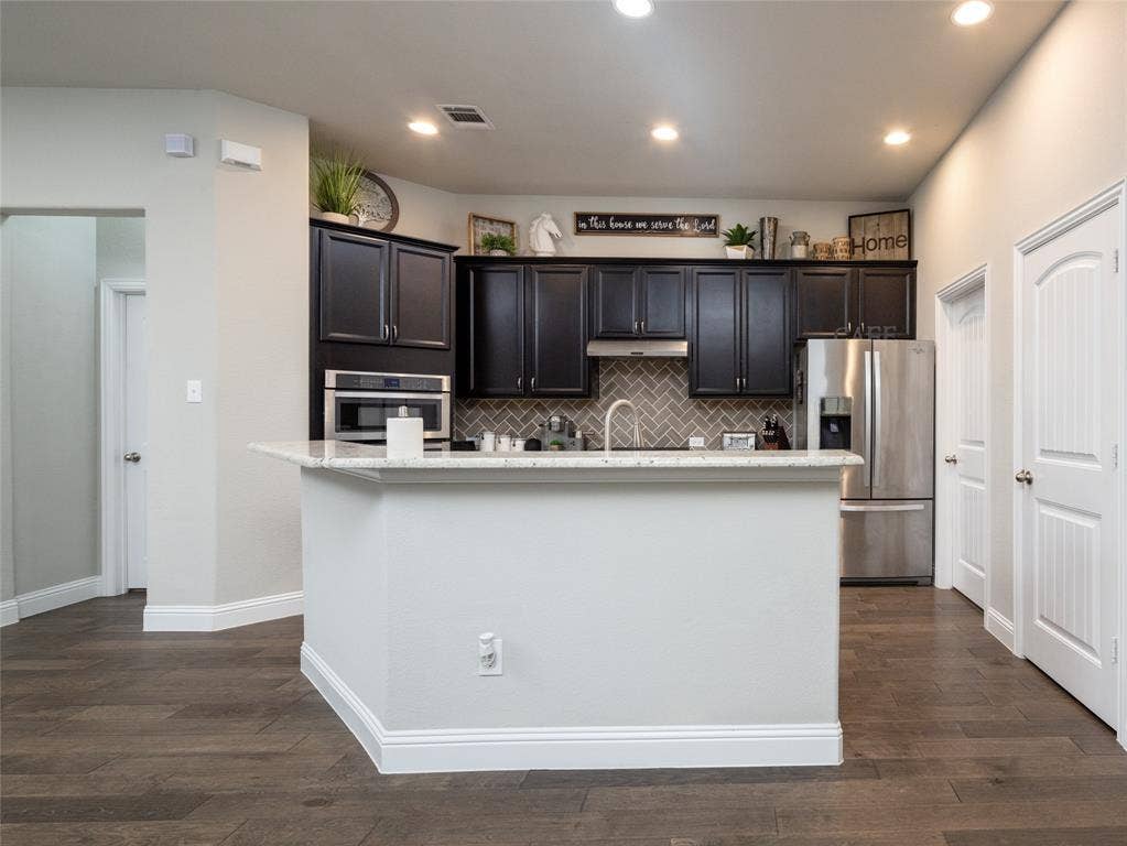 Kitchen island with a light-toned countertop, dark wood-finish cabinetry, stainless steel appliances, and a herringbone-patterned backsplash