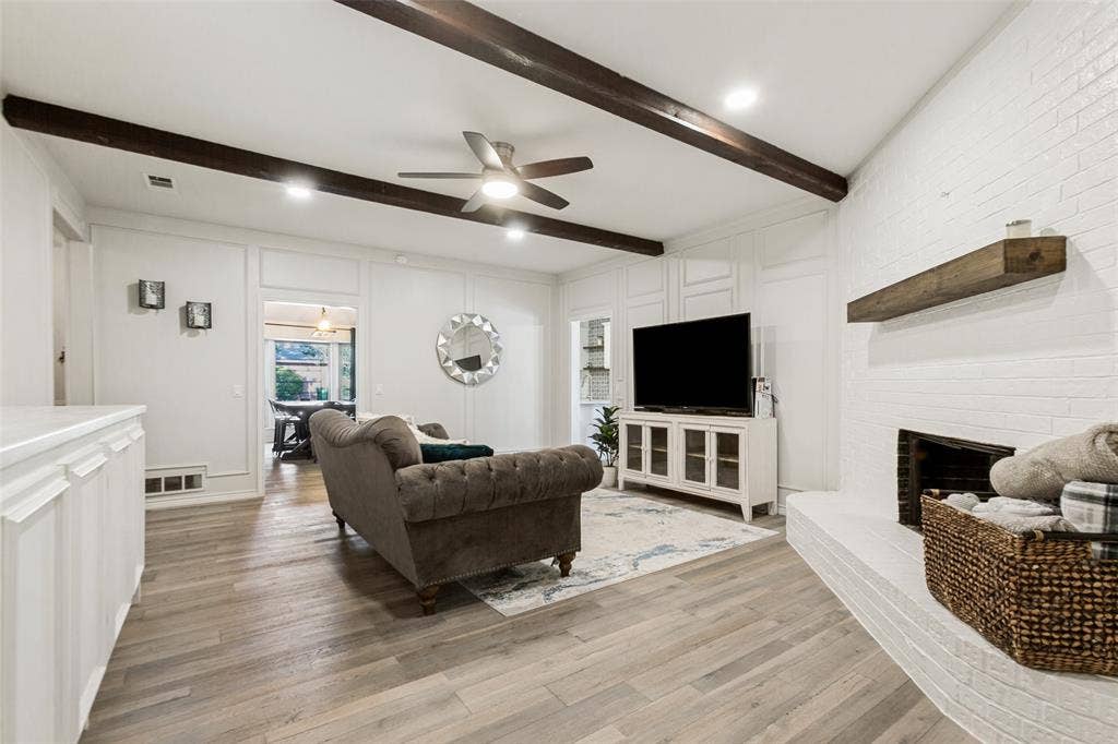 Wood-finish flooring, white brick fireplace with a wood mantle, white paneled wall accents, recessed lighting, and exposed ceiling beams