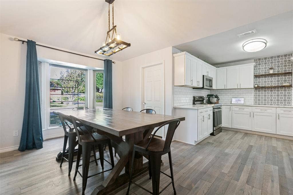 Open-concept kitchen and dining area featuring white cabinetry with stainless steel appliances, patterned tile backsplash, wood-finish flooring throughout, and large windows with blue drapery overlooking landscaped grounds