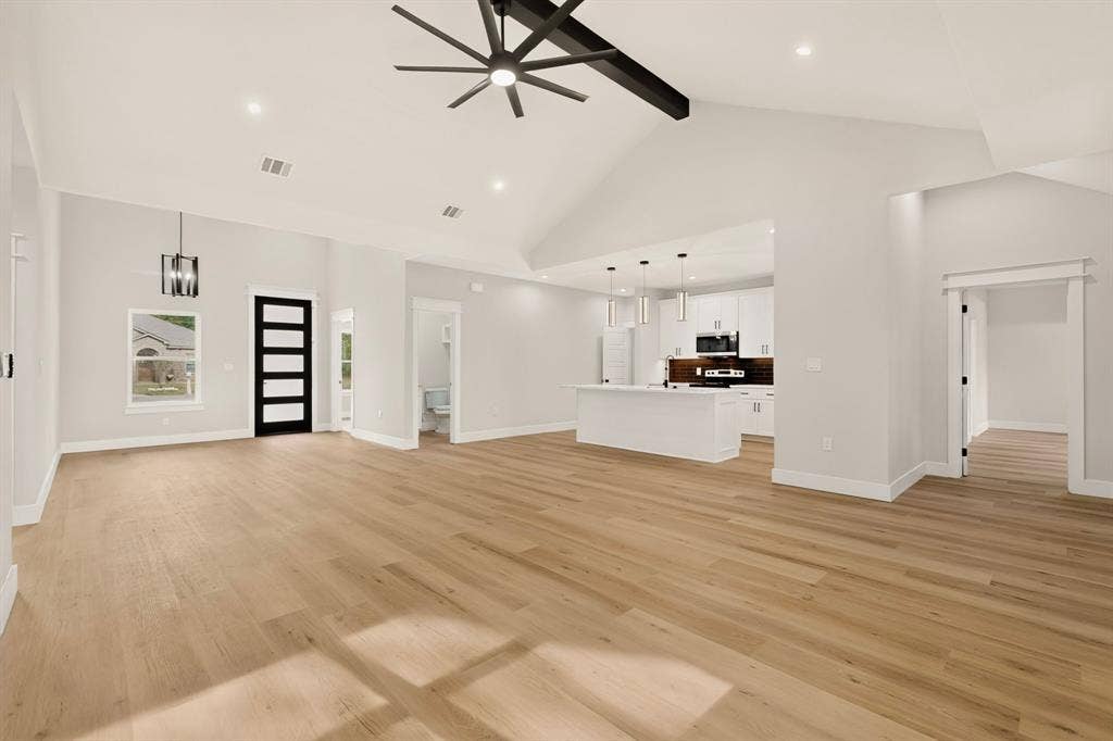 Spacious open-concept living area featuring high vaulted ceilings, light wood-finish flooring, a modern kitchen with a large island, and a distinctive black front entry door