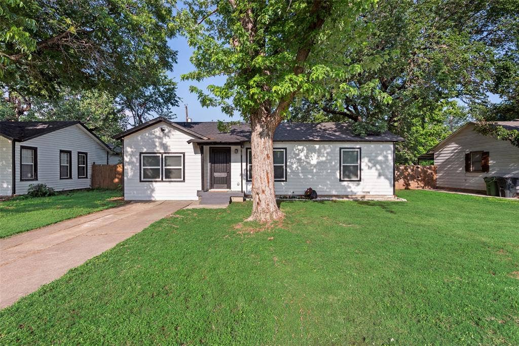 228 Harrison Lane, Hurst, TX 76053 White siding exterior with black trim windows, a dark shingled roof, a concrete driveway, and a large front yard with mature trees