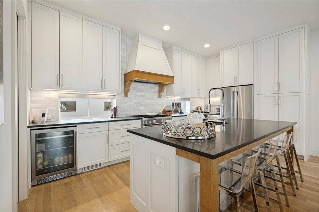 White shaker cabinetry, light wood-finish flooring, dark countertop island with seating, stainless steel appliances, and a built-in beverage refrigerator