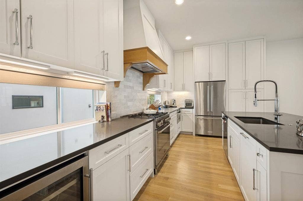 White cabinetry with brushed nickel hardware, stainless steel appliances, dark countertops, wood-finish flooring, and a kitchen island with an integrated sink