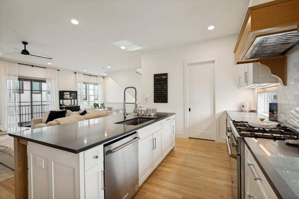 Open-concept kitchen featuring an island with a stainless steel sink, wood-finish flooring, a stainless steel range with a custom wood hood, and white cabinetry