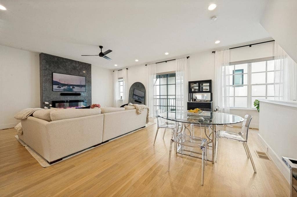 Open-concept living space featuring warm wood-finish flooring throughout, a cream upholstered sofa with modern lines, recessed ceiling lighting, and a ceiling fan