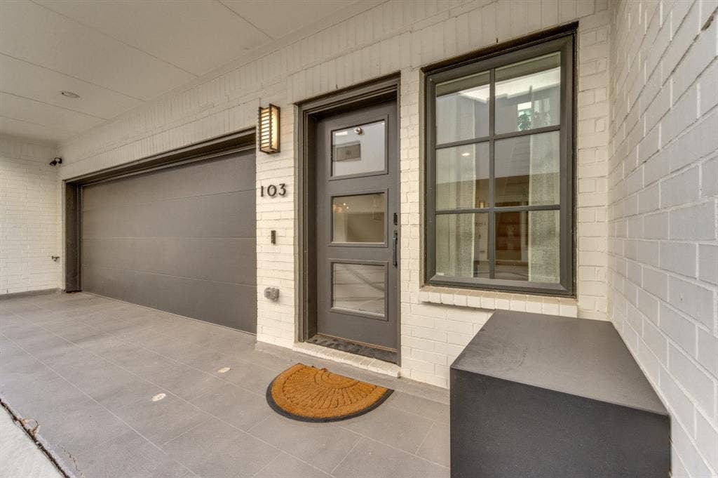 White brick exterior with a dark gray garage door, modern entry door with glass panels, and an exterior wall sconce