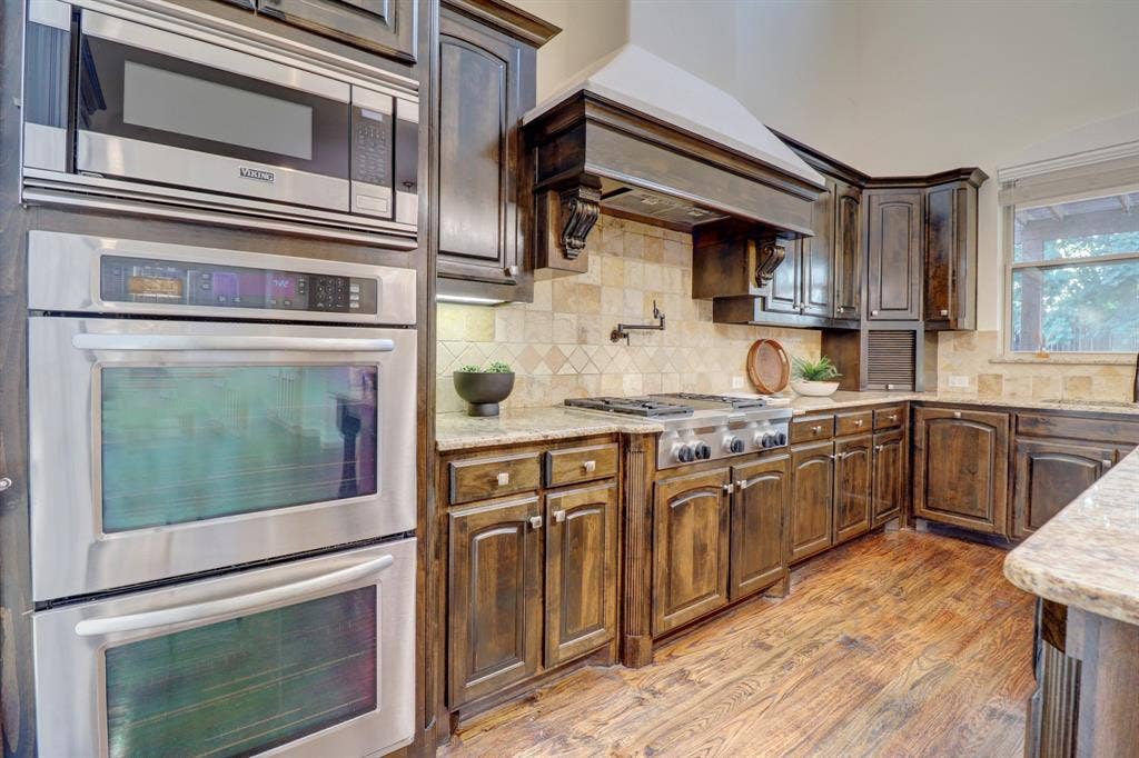 11559 Ashley Lane, Frisco, TX 75035 Dark wood-finish cabinetry, stainless steel double ovens, gas range with an ornate hood, tile backsplash, and light-toned countertops
