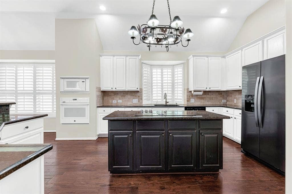 3217 Royal Melbourne Drive, Plano, TX 75093 A central dark island featuring a smooth cooktop serves as the hub, framed by crisp white cabinetry, stone tile backsplash, and a statement wrought-iron chandelier. You will love cooking family meals here with everything you need right at your fingertips.