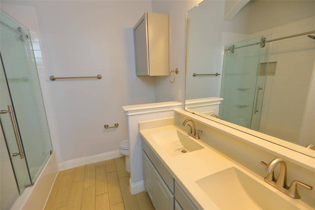 3707 Plum Vista Place, Arlington, TX 76005 Bathroom featuring a dual vanity with integrated sinks and brushed nickel faucets, a full-width mirror, light wood-finish flooring, and a glass-enclosed shower/tub combination