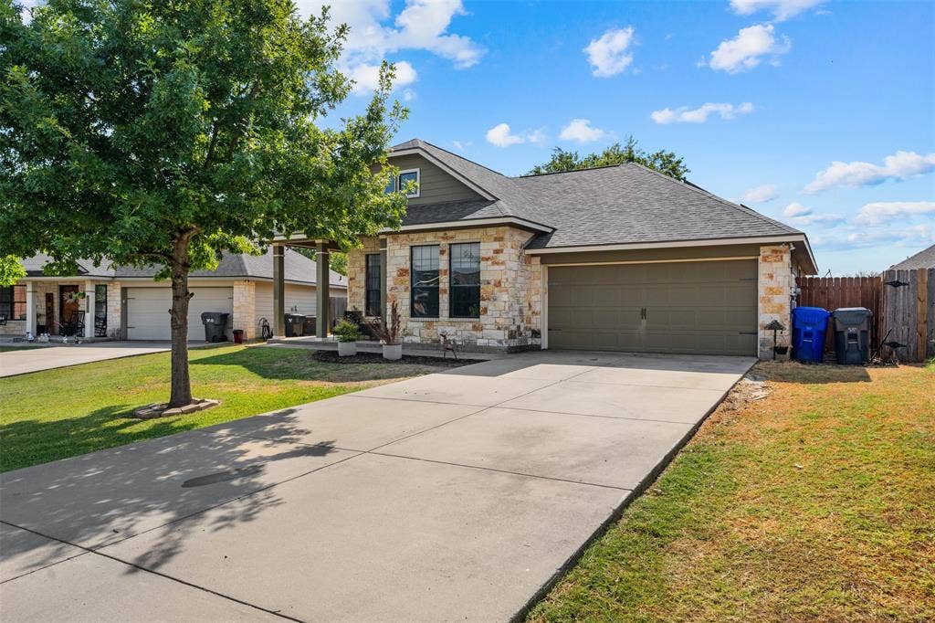 10313 Condor Loop, Waco, TX 76708 Stone veneer exterior with dark shingle roof, attached garage with a solid door, concrete driveway, and a front yard with mature trees