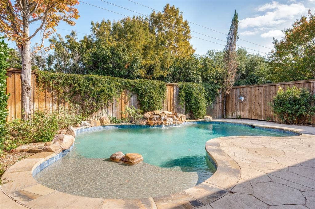 Custom-shaped swimming pool with a rock waterfall feature, integrated sun shelf, and natural stone coping—the fence has been recently stained, which isn't reflected in either backyard picture.