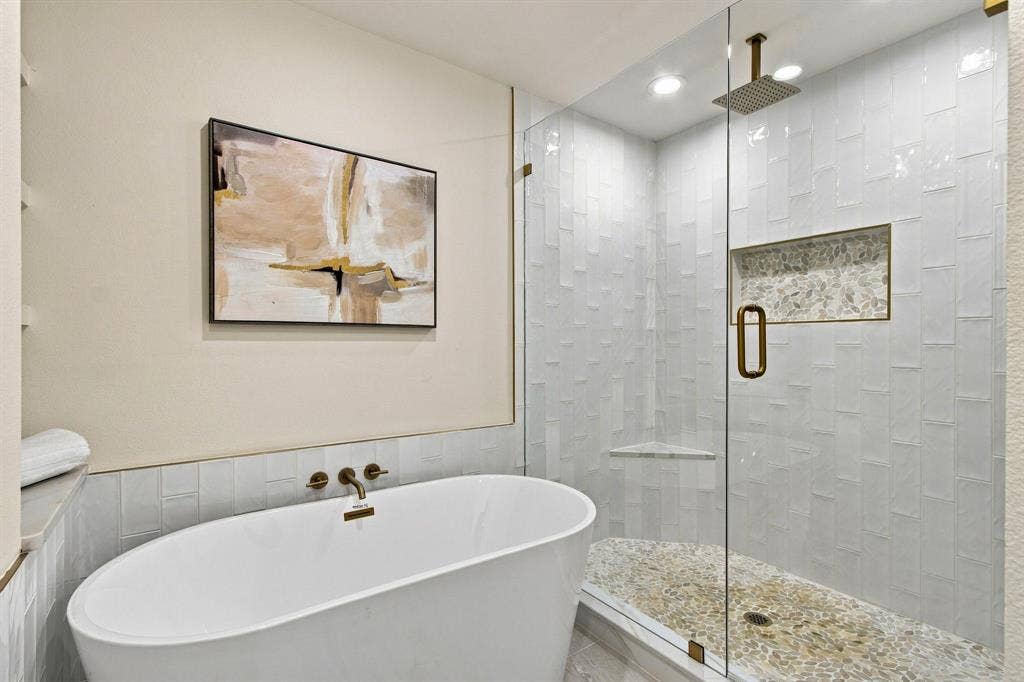 1409 Navaho Trail, Richardson, TX 75080 Freestanding soaking tub with wall-mounted bronze faucet, glass-enclosed shower with subway tile surround, recessed shower niche, and pebble-finish flooring, complemented by a ceiling-mounted bronze rain shower head