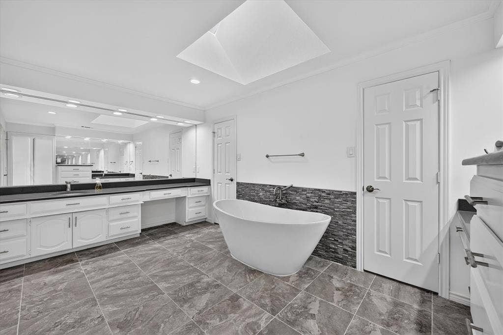 Freestanding soaking tub, skylight, extensive vanity with dual sinks, dark stone countertops, and large format floor tiling